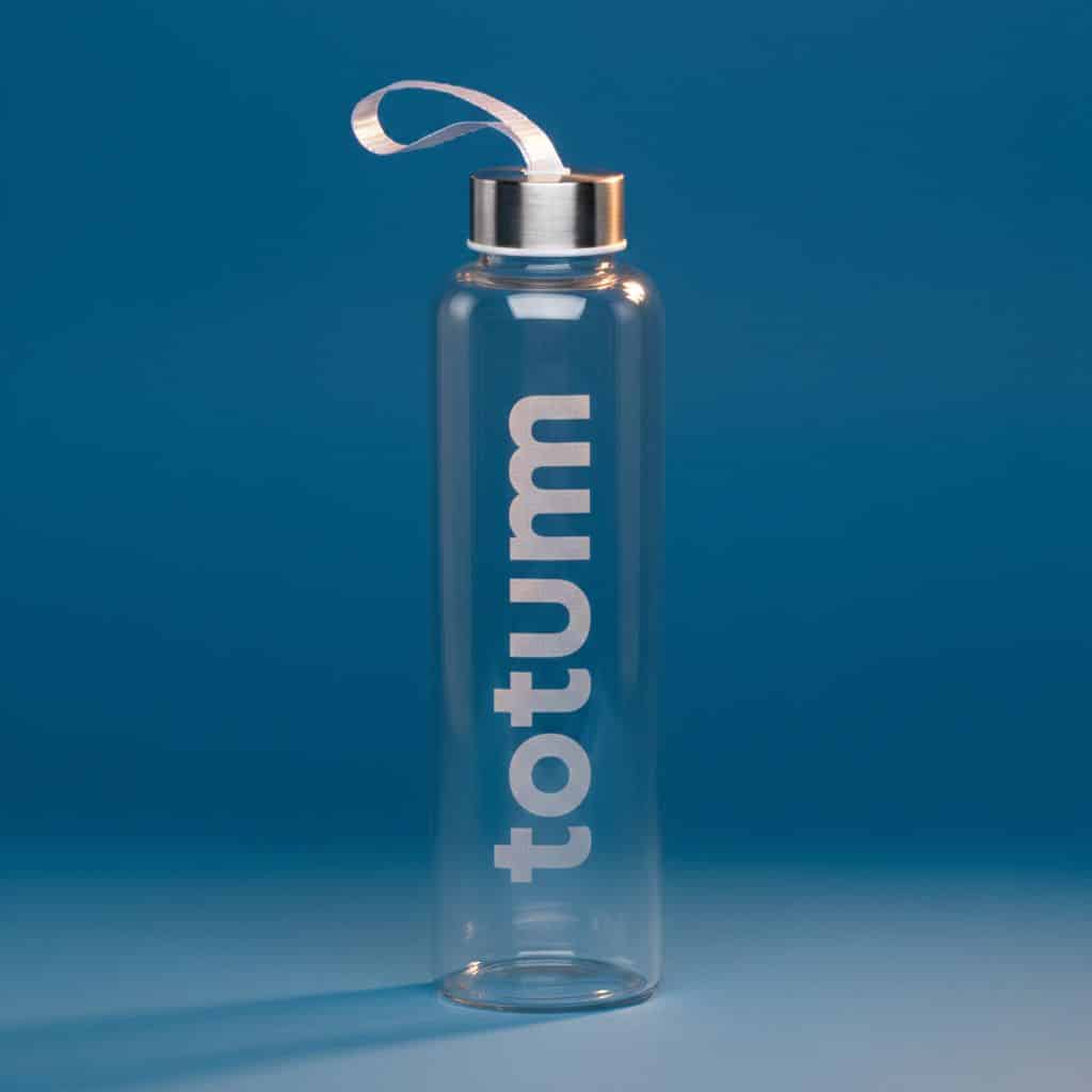 A clear glass water bottle with a silver screw-on lid and a carrying strap, standing upright on a blue surface with "totum" written vertically in white letters on the bottle.